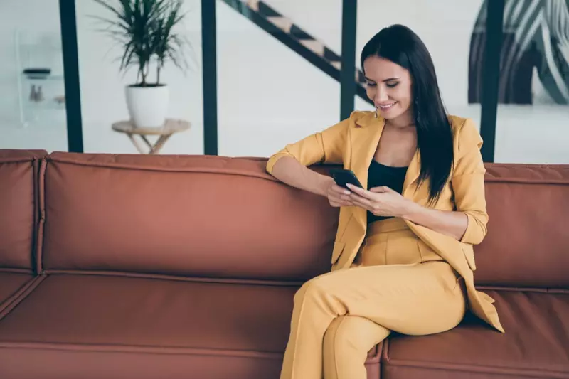 Žena v žltom nohavičkovom obleku na pohovke pri pohľade na telefón