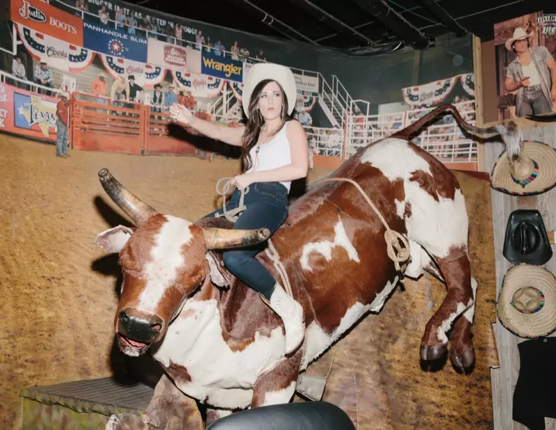 Kacey rider på en mekanisk tjur medan hon har en cowboyhatt, jeansbyxor och stövlar i västerländsk stil