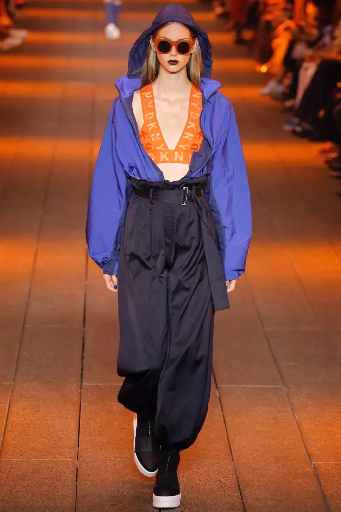 DKNY Primavera 2017: Lauren de Graaf camina per la passarel·la amb una jaqueta amb caputxa de niló flotant de color blau pilot i la clàssica jaqueta de niló flotant, un sostenidor amb logotip taronja i un pantaló clàssic de bossa de paper de setí marí.