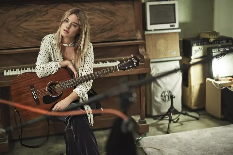 Camille Rowe posiert mit einer Gitarre und trägt eine Bluse mit Schleifenmuster und eine Samthose